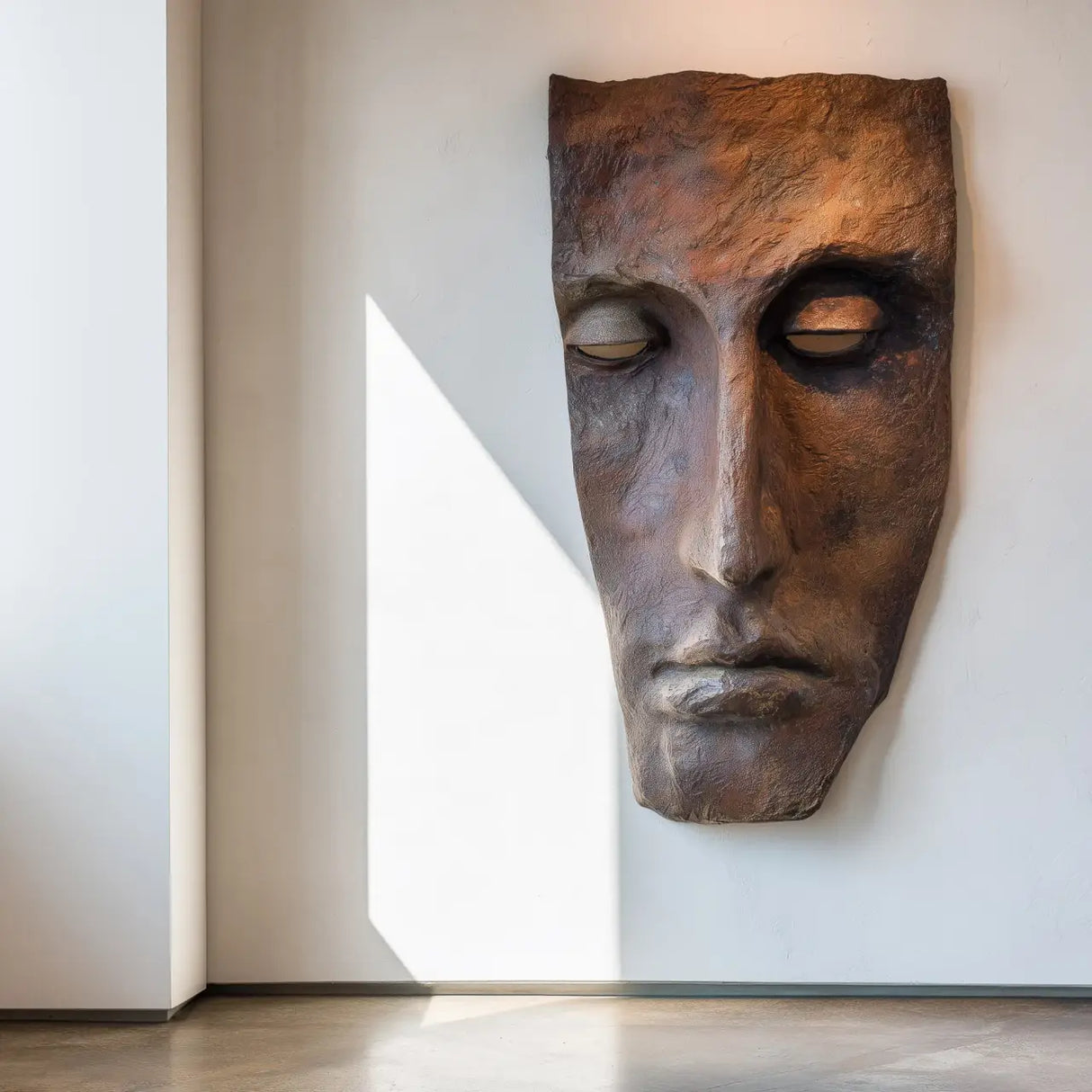 A realistic bronze wall mask sculpture in human face design is mounted on a plain wall in a sunlit modern interior.