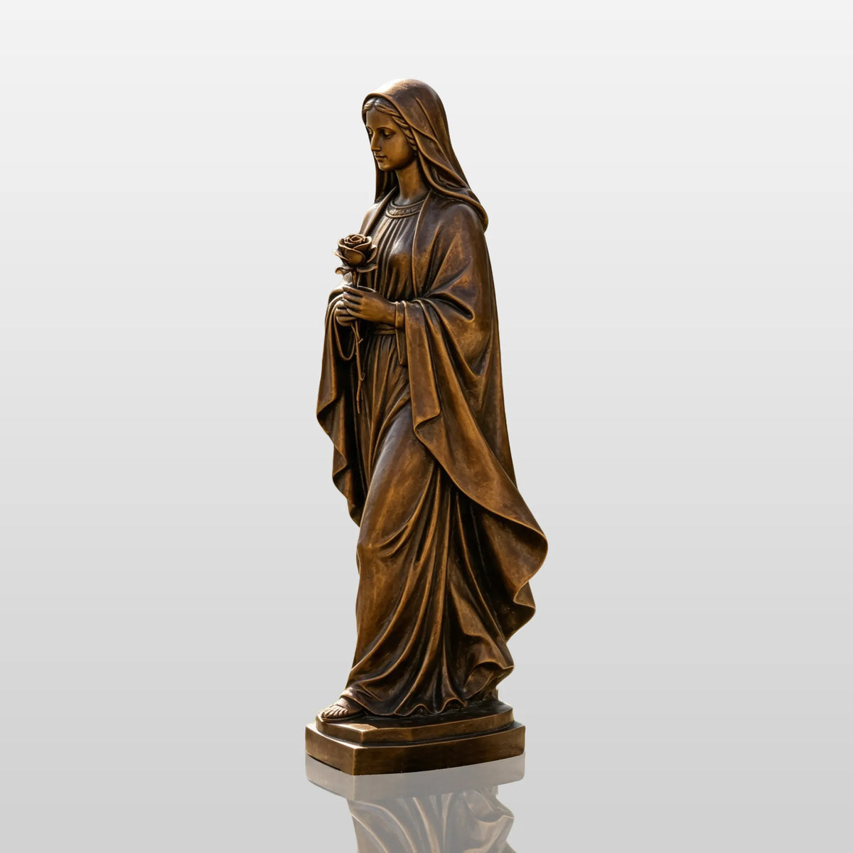 A bronze Virgin Mary statue stands on the grass beside the sidewalk in front of a residential house.