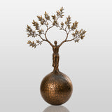 Bronze sculpture with the design of human shaped tree ball stands in a cobblestone courtyard.
