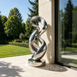 Stainless steel twisted loop sculpture stands on the outdoor patio beside a modern building.