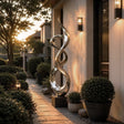 An abstract twisted stainless steel sculpture stands beside a walkway at sunset amid trimmed green shrubs.