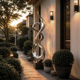 An abstract twisted stainless steel sculpture stands beside a walkway at sunset amid trimmed green shrubs.