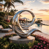 Mirror wave stainless steel sculpture sits besides ocean with palm trees at sunset.