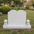 A white marble tombstone with two puppy statues stands on green grass inside a cemetery.