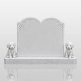 A white marble tombstone with two puppy statues stands on green grass inside a cemetery.