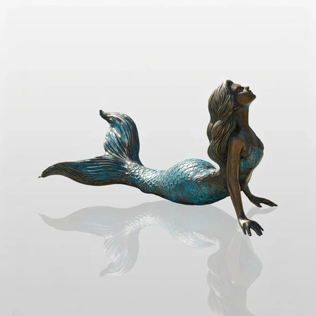 A bronze mermaid sculpture rests on a pool coping beside clear blue pool water surrounded by tropical blooming shrubs.