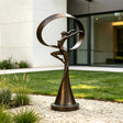 The bronze dancer sculpture with a ribbon sits on gravel beside green lawn.