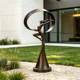 The bronze dancer sculpture with a ribbon sits on gravel beside green lawn.