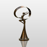 The bronze dancer sculpture with a ribbon sits on gravel beside green lawn.