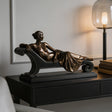 Bronze tabletop reclining lady sculpture is displayed on black cabinet within modern styled interior space.