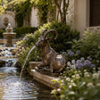 Antique bronze ibex fountain spouts water into garden pond surrounded by blooms, trimmed shrubs.