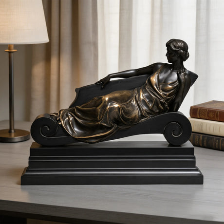 Bronze reclining lady table sculpture sits on light indoor table beside vintage books and table lamp.