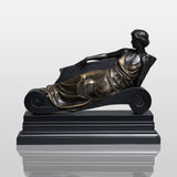 Bronze reclining lady table sculpture sits on light indoor table beside vintage books and table lamp.