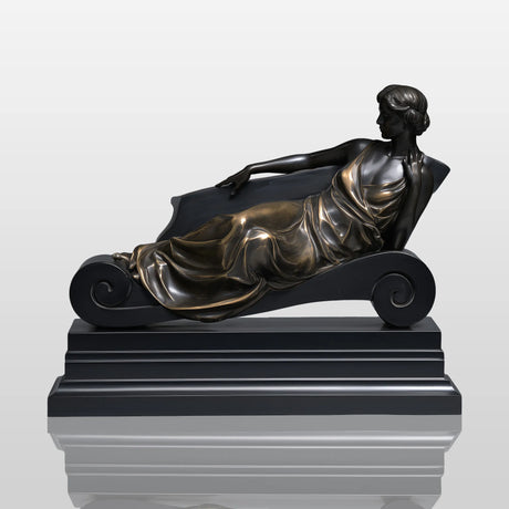 Bronze reclining lady table sculpture sits on light indoor table beside vintage books and table lamp.