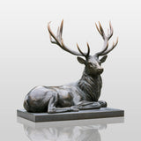 Bronze deer statue rests on a flat base within a lush green park.