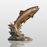 A bronze leaping salmon sculpture rests on rocks beside a lake with mountains and trees in the background.