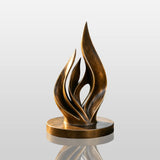 Bronze abstract flame sculpture stands under sunshine inside a green garden.