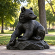 Dark bronze seated bear statue sits on pavement within a green public park.