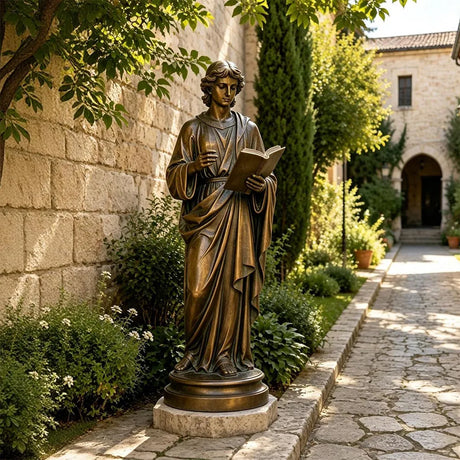 A bronze St. John the Apostle statue stands on stone paving in a sunlit courtyard garden.