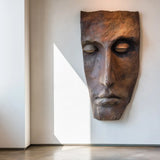 A realistic bronze wall mask sculpture in human face design is mounted on a plain wall in a sunlit modern interior.