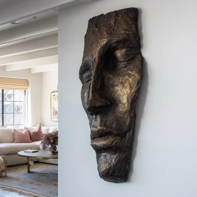 A modern bronze face wall mask sculpture hangs on a light colored wall in a bright, contemporary living room.