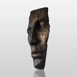 A modern bronze face wall mask sculpture hangs on a light colored wall in a bright, contemporary living room.