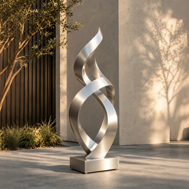 A stainless steel twist sculpture stands on paved ground outside a modern building.