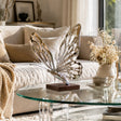 A hollow polished stainless steel butterfly sculpture sits on a curved glass table inside a bright living room.