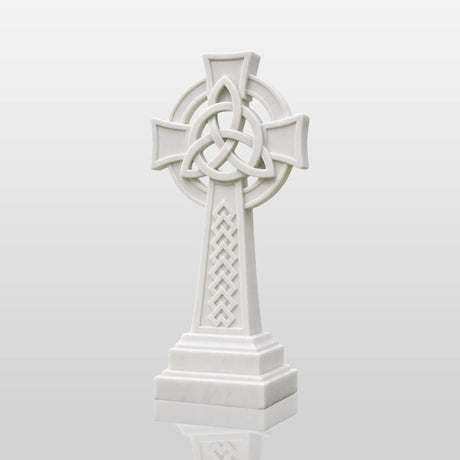 White marble Celtic cross headstone rests on gravel ground surrounded.