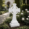 A marble Celtic cross tombstone with triangle knots sits among blooming roses near a church.