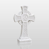 A marble Celtic cross tombstone with triangle knots sits among blooming roses near a church.