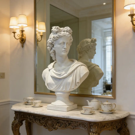 A white Greek Apollo marble bust sits on a carved marble console table before a gold framed mirror in a classic interior.