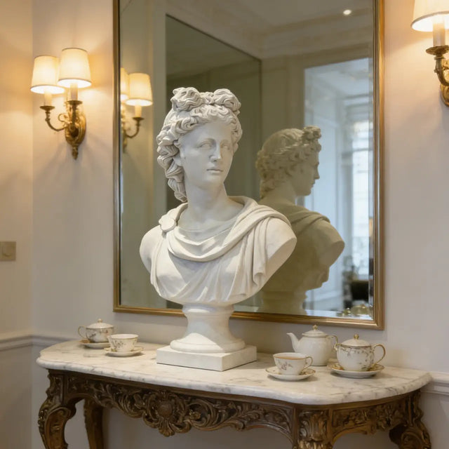 A white Greek Apollo marble bust sits on a carved marble console table before a gold framed mirror in a classic interior.