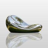 A mirror polished organic curved stainless steel bench rests on lush lawn under sun.