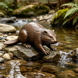 Bronze beaver statue crouches on wet rocks beside a flowing forest stream in a garden.