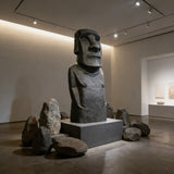 A large textured Moai stone statue rests surrounded by natural rocks inside a minimalist art gallery lit by ceiling spotlights.