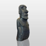 A large textured Moai stone statue rests surrounded by natural rocks inside a minimalist art gallery lit by ceiling spotlights.