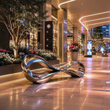 Mirror polished abstract stainless steel bench sculpture rests on tiled mall walkway beside flower planters and glowing light strips at night.