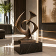 A bronze sculpture with intertwined shapes rests on a dark stone base inside a room.