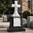 White ivy marble cross headstone with blank scroll plaque sits on black granite base amid gray tombstones in a sunlit cemetery.