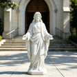 A white marble Jesus statue with open hands stands on the pavement in front of a church entrance.
