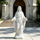 A white marble Jesus statue with open hands stands on the pavement in front of a church entrance.