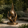 A bronze layered flame sculpture rests on stone walkway amid lush garden plants.