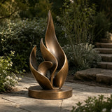 A bronze layered flame sculpture rests on stone walkway amid lush garden plants.