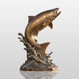 A bronze leaping salmon statue with wave base sits on the ground in front of lake and mountains.
