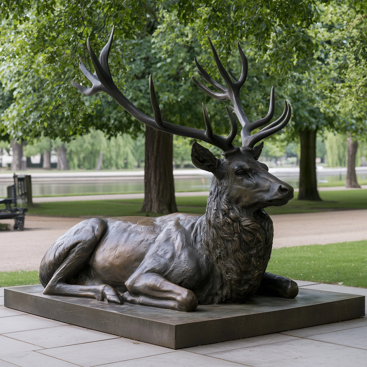 Bronze lying deer statue sits on stone plinth amid green park surroundings.