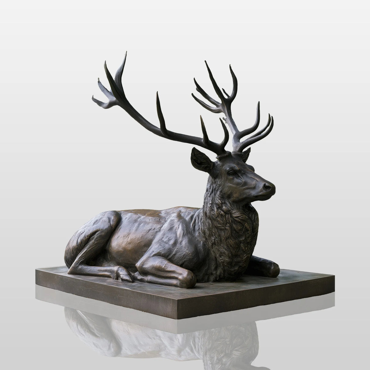 Bronze lying deer statue sits on stone plinth amid green park surroundings.