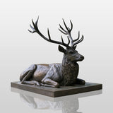 Bronze lying deer statue sits on stone plinth amid green park surroundings.