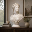 A white classical marble bust statue sits on a dark wooden sideboard beside stacked books and small potted greenery in a soft lit interior room.