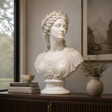 A white classical marble bust statue sits on a dark wooden sideboard beside stacked books and small potted greenery in a soft lit interior room.